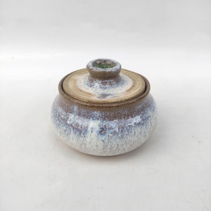 May include: A ceramic sugar bowl with a lid. The bowl is round with a brown rim and a white and blue glaze. The lid has a small knob and a brown rim. The sugar bowl is a decorative kitchen item.