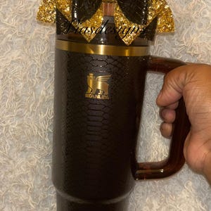 May include: A black and gold Stanley tumbler with a decorative bow. The tumbler has a textured, snake-skin-like pattern and a gold rim. The bow is black with gold glitter accents. The handle is a dark brown color.