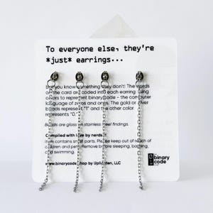 Może przedstawiać: Cztery srebrne kolczyki łańcuszkowe przymocowane do białej karty ekspozycyjnej. Karta zawiera tekst: To everyone else, they're *just* earrings... But you know something they don't! The words on the card are coded into each earring using colors to represent binary code - the computer language of zeros and ones. The gold or silver beads represent '1' and the other color represents '0.' Beads are glass on stainless steel findings. Compiled with love by nerds. Item contains small parts. Please keep out of reach of children and pets. Remove before sleeping, bathing, and swimming. The binary code shop by Uplinten, LLC.