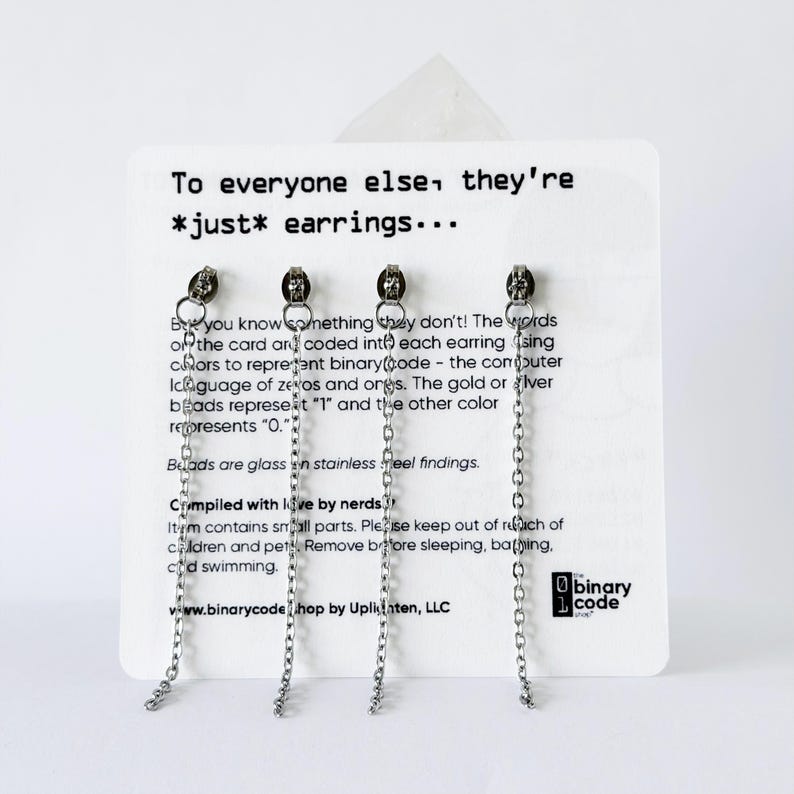 Może przedstawiać: Cztery srebrne kolczyki łańcuszkowe przymocowane do białej karty ekspozycyjnej. Karta zawiera tekst: To everyone else, they're *just* earrings... But you know something they don't! The words on the card are coded into each earring using colors to represent binary code - the computer language of zeros and ones. The gold or silver beads represent '1' and the other color represents '0.' Beads are glass on stainless steel findings. Compiled with love by nerds. Item contains small parts. Please keep out of reach of children and pets. Remove before sleeping, bathing, and swimming. The binary code shop by Uplinten, LLC.