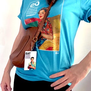 Peut inclure: Un t-shirt bleu vif avec un graphique repr&eacute;sentant une personne en chemise &agrave; fleurs et du code binaire indiquant GO YOURSELF. Un sac bandouli&egrave;re en cuir marron est port&eacute; &agrave; l'&eacute;paule, orn&eacute; d'une &eacute;tiquette rectangulaire montrant une personne et EAT.