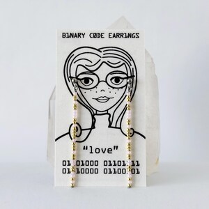 May include: A pair of long, linear earrings featuring alternating gold and light pink beads, displayed on a white card labeled BINARY CODE EARRINGS with a line drawing of a person wearing glasses and the text love and binary code sequences.
