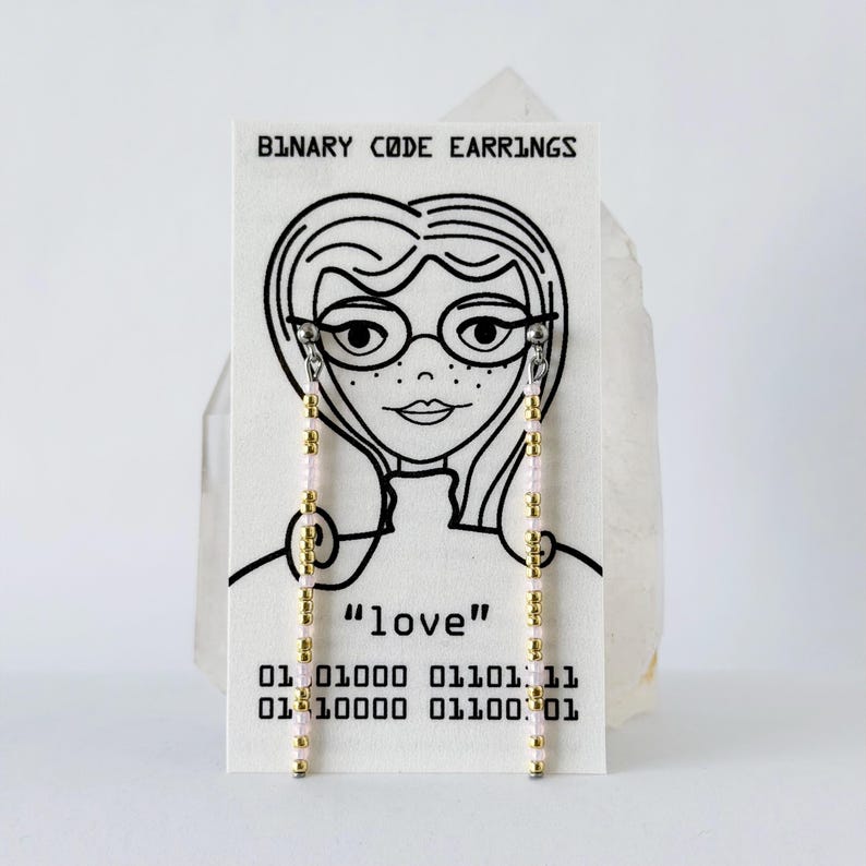 Może przedstawiać: Para długich, liniowych kolczyk&oacute;w z naprzemiennymi złotymi i jasnor&oacute;żowymi koralikami, prezentowanych na białej karcie z napisem BINARY CODE EARRINGS, rysunkiem liniowym osoby w okularach, słowem love oraz sekwencjami kodu binarnego.
