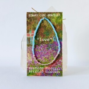 May include: A beaded bracelet featuring light blue and pink beads on a rectangular display card. The card displays the text BINARY CODE BRACELET, the word love in quotes, and a sequence of binary numbers in white text over a floral garden image.