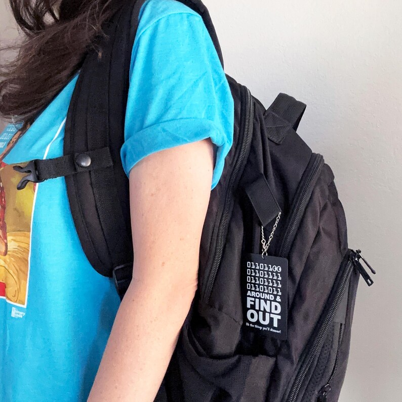 Peut inclure: Un porte-cl&eacute;s rectangulaire noir attach&eacute; &agrave; un sac &agrave; dos noir par une cha&icirc;ne argent&eacute;e. Le porte-cl&eacute;s pr&eacute;sente du code binaire blanc en haut, le texte "AROUND & FIND OUT" en grandes lettres blanches et le texte "Oh the things you'll discover!" en dessous.