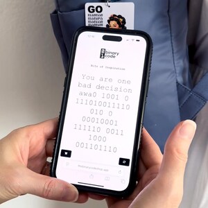 Peut inclure: Un smartphone affichant un site web avec le texte "binary code" et "Bits of Inspiration" suivi de "You are one bad decision awa0" et des s&eacute;quences binaires. Une &eacute;tiquette de bagage blanche avec "GO" et du texte binaire pend d'un sac &agrave; dos bleu.
