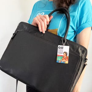Peut inclure: Une mallette rectangulaire noire avec deux poign&eacute;es sup&eacute;rieures et une poche frontale zipp&eacute;e. Une &eacute;tiquette de bagage rectangulaire est suspendue &agrave; la poign&eacute;e, affichant le texte GO, du code binaire et une illustration color&eacute;e d'une personne en chemise rouge.