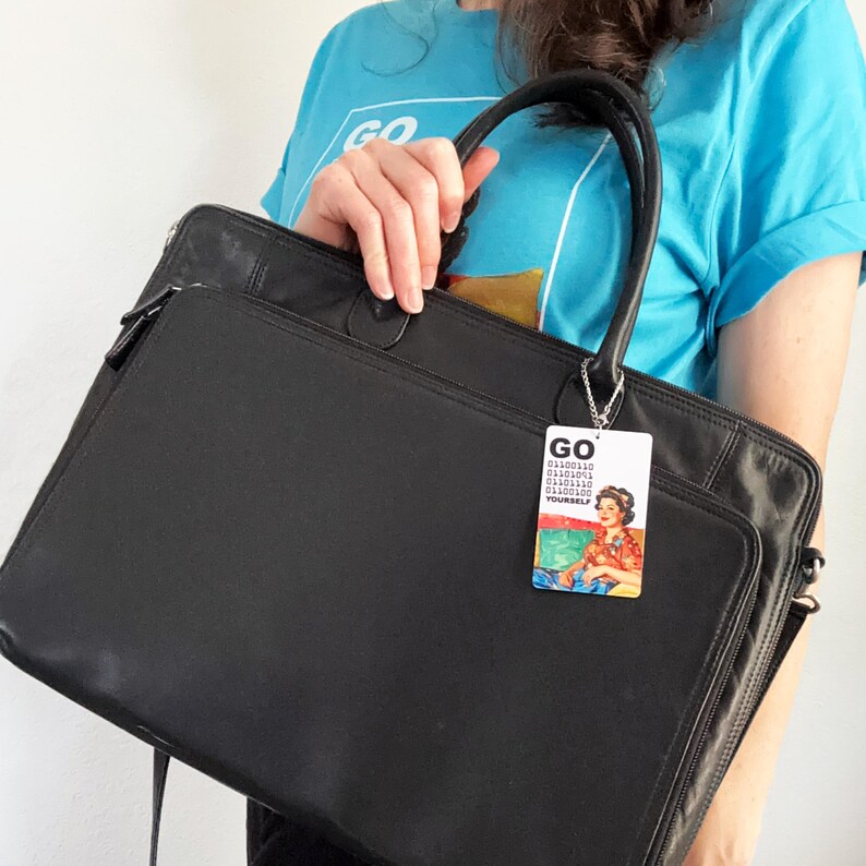 Peut inclure: Une mallette rectangulaire noire avec deux poign&eacute;es sup&eacute;rieures et une poche frontale zipp&eacute;e. Une &eacute;tiquette de bagage rectangulaire est suspendue &agrave; la poign&eacute;e, affichant le texte GO, du code binaire et une illustration color&eacute;e d'une personne en chemise rouge.