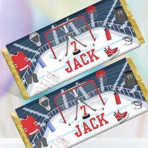 Peut inclure: Deux emballages de barres chocolatées sur le thème du hockey. Chaque emballage présente un design de patinoire avec un filet, des crosses et le nom "JACK" en rouge. Les emballages sont conçus pour des barres chocolatées de 44 g.