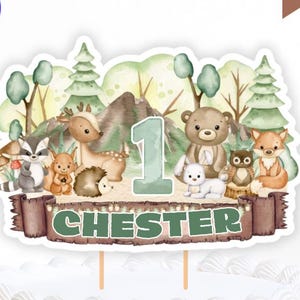 Op de afbeelding: Een taarttopper met bosdieren met een bosscène, met een hert, beer, vos en andere dieren. Het nummer 1 wordt weergegeven, samen met de naam "Chester". De taarttopper staat op een wit geglazuurde taart. De woorden "Digitale download" zijn zichtbaar.