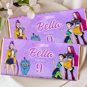 Puede incluir: Dos envoltorios de barras de chocolate con fondo morado y bordes dorados. Los envoltorios presentan ilustraciones de personajes de dibujos animados y el texto "Bella turns 9". Los envoltorios miden 13,3 x 14 cm.