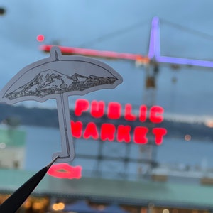 May include: A black and white sticker of an umbrella with a mountain inside. The sticker is placed on a window with a red neon sign that says "PUBLIC MARKET" in the background.