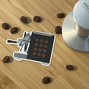 以下が含まれることがあります： 黒い前面と茶色のドット模様のエスプレッソマシンステッカー。コーヒー豆とシルバーのRocket Espresso Milanoタンパーが竹の表面にあります。真上からの画像です。