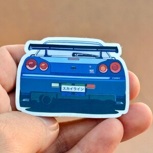 Op de afbeelding: Blauwe autosticker met de achterkant van een sportwagen. De sticker heeft een zwarte spoiler en Japanse tekst op de nummerplaat. De achterlichten van de auto zijn rood. De sticker wordt door een hand vastgehouden.