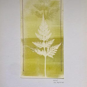 May include: A botanical print featuring a white fern silhouette against a yellow-green background. The artwork is framed with a black dotted line and includes the text "upright" and the artist's signature. The print is on a rectangular piece of paper.