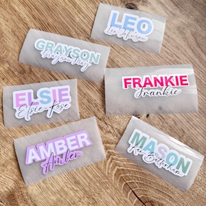 May include: A selection of personalised name stickers in shades of blue, pink, and green. Each sticker displays a name in a bold, colourful font, with a smaller, cursive font beneath. The stickers are on a clear backing.