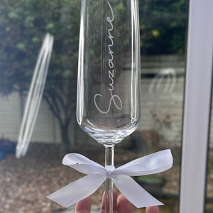 May include: A clear glass champagne flute with the name "Suzanne" etched on the side. A white satin ribbon is tied around the stem of the glass. The base of the glass has the number "50th" etched on it.