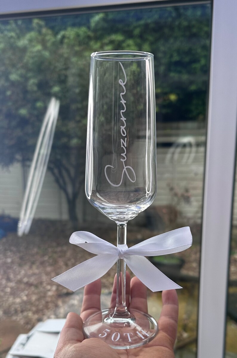 May include: A clear glass champagne flute with the name "Suzanne" etched on the side. A white satin ribbon is tied around the stem of the glass. The base of the glass has the number "50th" etched on it.