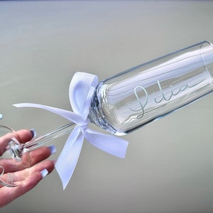 May include: A clear glass champagne flute with a white ribbon tied around the stem. The flute is engraved with the name "Julia" and the base of the stem is engraved with the word "PROM".