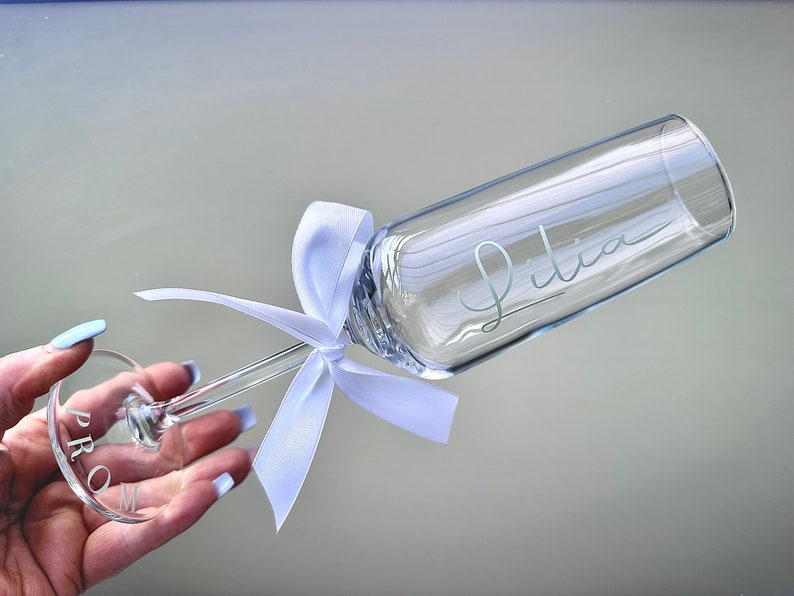 May include: A clear glass champagne flute with a white ribbon tied around the stem. The flute is engraved with the name "Julia" and the base of the stem is engraved with the word "PROM".
