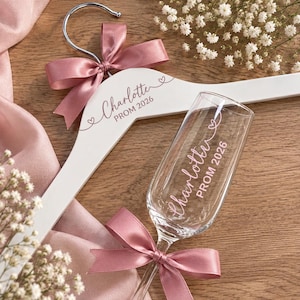 May include: A white wooden clothes hanger and a clear glass champagne flute. The hanger features a dusty pink satin bow and the text Charlotte PROM 2026. The flute is decorated with a matching pink satin bow and the text Charlotte PROM 2026.