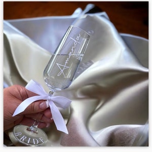 May include: A champagne flute with the name "Amanda" etched on the side. The flute is filled with champagne and has a white ribbon tied around the stem. The base of the flute says "Bride".
