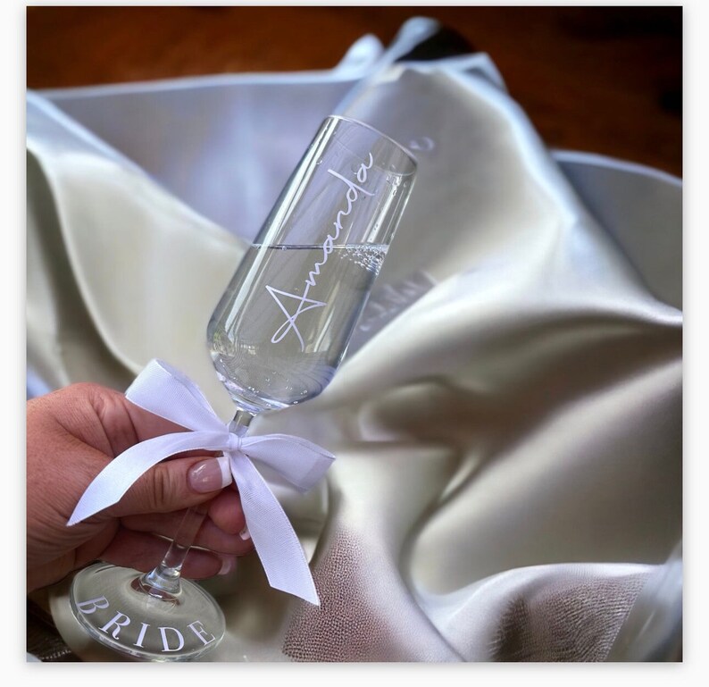 May include: A champagne flute with the name "Amanda" etched on the side. The flute is filled with champagne and has a white ribbon tied around the stem. The base of the flute says "Bride".
