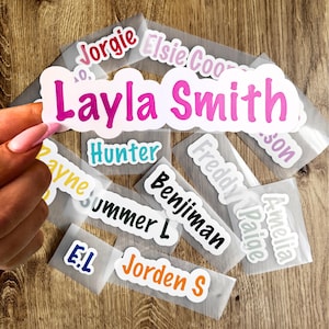 May include: A selection of name stickers in diverse colours and fonts, including "Layla Smith," "Hunter," and "Jorden S." The stickers are on transparent backing, ready for use. These are suitable for personalizing belongings.