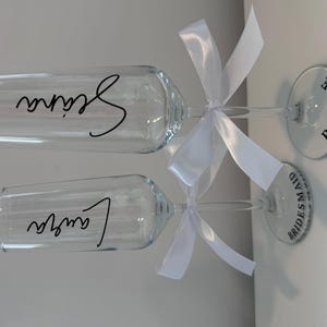 May include: Two champagne flutes with white bows and personalized labels. The labels read "Bride" and "Bridesmaid".
