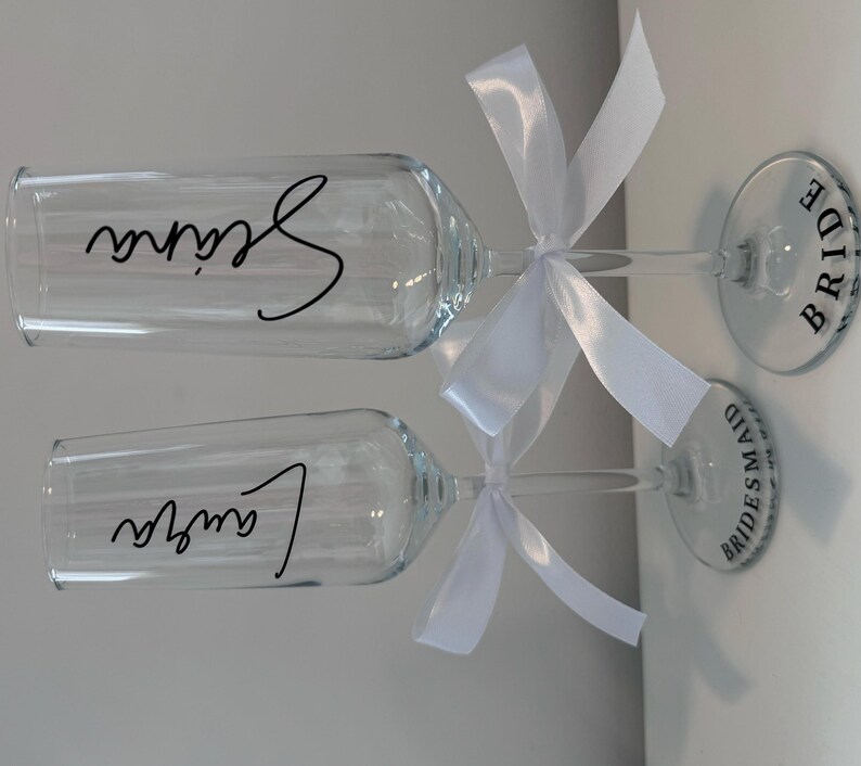 May include: Two champagne flutes with white bows and personalized labels. The labels read "Bride" and "Bridesmaid".