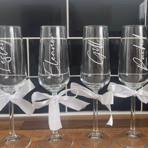 May include: Four clear glass champagne flutes with white satin ribbons tied around the stems. Each flute is personalized with a name: Lesley, Eleanor, Gill, and Rachel.