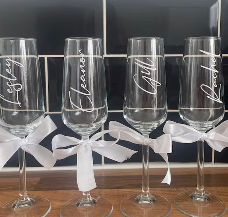May include: Four clear glass champagne flutes with white satin ribbons tied around the stems. Each flute is personalized with a name: Lesley, Eleanor, Gill, and Rachel.