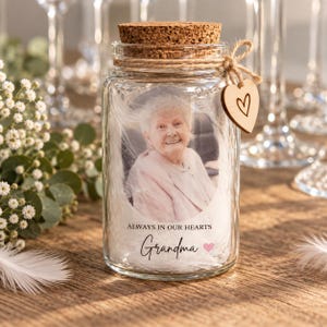 May include: A clear glass jar with a cork stopper, filled with white feathers and a photograph of an elderly person. The jar features the text 'ALWAYS IN OUR HEARTS' and 'Grandma' with a small pink heart, plus a wooden heart tag tied with twine.