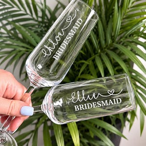 May include: Two clear glass champagne flutes featuring white custom lettering. One flute is personalised with the name Chloe and the word BRIDESMAID, while the other flute is personalised with the name Ellie and the word BRIDESMAID, both with a heart.