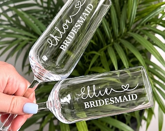 May include: Two clear glass champagne flutes featuring white custom lettering. One flute is personalized with the name Chloe and the word BRIDESMAID, while the other flute is personalized with the name Ellie and the word BRIDESMAID, both with a heart.