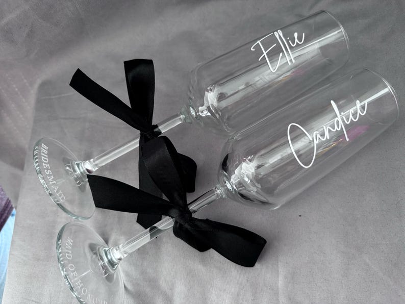 May include: Two champagne flutes with black ribbon bows. One flute says "Maid of Honor" and the other says "Bridesmaid".