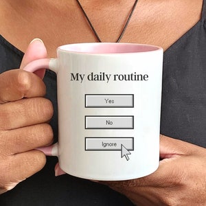 Könnte beinhalten: Eine weiße Keramiktasse mit rosafarbenem Rand und Henkel. Auf der Tasse steht der Text "My daily routine" gedruckt, darunter drei Knöpfe: "Yes", "No" und "Ignore". Ein Computermauszeiger zeigt auf den Knopf "Ignore".