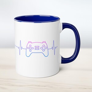 May include: White ceramic coffee mug with a blue rim and handle. The mug features a pink and blue outline of a video game controller with a heartbeat line.