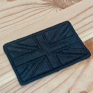 May include: A rectangular embroidered patch featuring a monochromatic black Union Jack flag design. The patch has a stitched border and a textured surface with raised thread detailing that forms the iconic cross and diagonal lines of the flag.