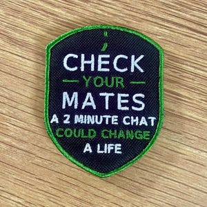 May include: A shield-shaped embroidered patch with a black background and a green border. The text reads: CHECK YOUR MATES, A 2 MINUTE CHAT COULD CHANGE A LIFE. The words CHECK, MATES, and A 2 MINUTE CHAT are in white, while YOUR and COULD CHANGE A LIFE are in green.