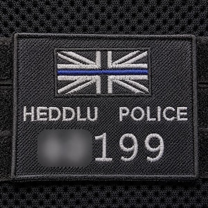 May include: A rectangular black embroidered patch featuring a stylized Union Jack flag with a thin horizontal blue line across the centre. The text HEDDLU POLICE is stitched in white above the number 199, with a blurred area to the left of the number.