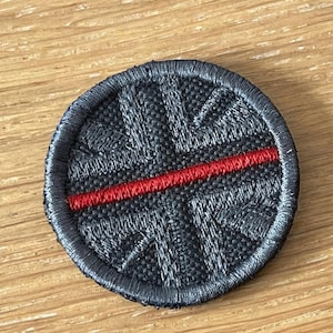 May include: A circular embroidered patch featuring a black and dark grey Union Jack flag design. A single horizontal red line is stitched across the centre of the patch, which has a thick, dark grey stitched border around the entire circumference.