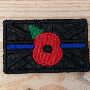 May include: A rectangular embroidered patch featuring a black Union Jack design with a thin horizontal blue line running through the centre. A bright red poppy flower with a black centre and a small green leaf is stitched over the middle section.