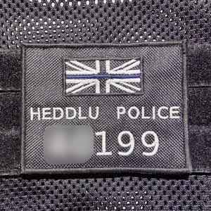 May include: A black patch displaying the words "HEDDLU POLICE" and the number "199" in white. The patch includes a Union Jack flag with a blue line. The patch is attached to a black mesh material.