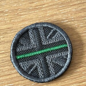 May include: A circular embroidered patch featuring a black and dark grey Union Jack flag design. A single horizontal line of bright green thread runs across the centre of the patch, dividing the flag pattern. The edges are finished with a black border.