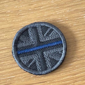 May include: A circular embroidered patch featuring a dark grey Union Jack flag design. A single horizontal blue line runs across the centre of the patch, dividing the flag pattern. The edges of the circular patch are finished with a dark grey stitched border.