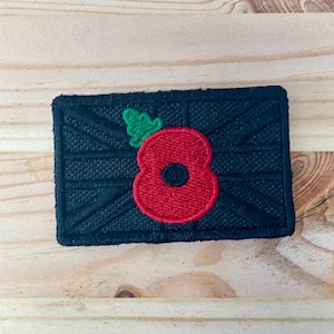 May include: A rectangular embroidered patch featuring a black Union Jack flag design as the background. Centered on the patch is a bright red poppy flower with a small green leaf attached to the top left side of the flower petals.