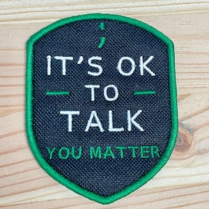 May include: A shield-shaped embroidered patch with a black background and a green border. It features a semicolon symbol, the white text "IT'S OK TO TALK" with green horizontal lines, and the green text "YOU MATTER" at the bottom of the patch.
