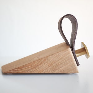 Könnte beinhalten: Ein Holz-Türstopper mit einem braunen Ledergriff und einem Messingknopf. Der Türstopper ist schräg und hat eine natürliche Holzmaserung.