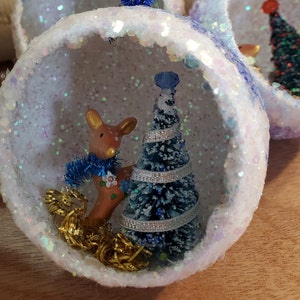 May include: A hollow, spherical ornament covered in white iridescent glitter containing a miniature brown deer figurine with a blue tinsel scarf, a small green bottle brush tree with silver ribbon, and gold metallic tinsel at the base.